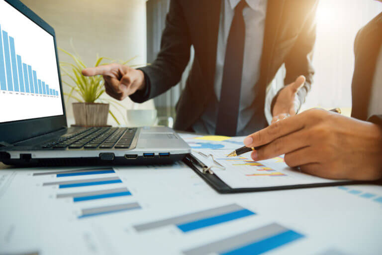 A business professional pointing at a laptop screen displaying a downward bar chart, while another person is holding a pen over a document with financial graphs and charts, symbolizing financial analysis or strategy discussions.