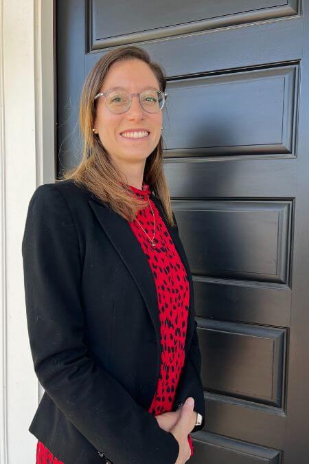 Maureen Donahue, CFP®, stands confidently at the entrance of Yardley Wealth Management, located in Yardley, Pennsylvania. She's wearing a black blazer with a red blouse, standing against a dark blue door with a white frame. Sporting glasses, her light brown hair frames her face, and she greets visitors with a big smile. As an esteemed employee of the firm, her expertise and dedication reflect the commitment to excellence upheld by Yardley Wealth Management.