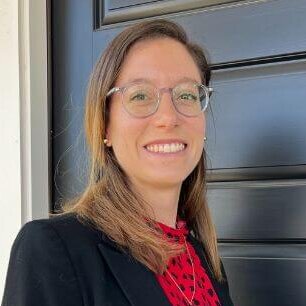 Maureen Donahue, CFP®, stands confidently at the entrance of Yardley Wealth Management, located in Yardley, Pennsylvania. She's wearing a black blazer with a red blouse, standing against a dark blue door with a white frame. Sporting glasses, her light brown hair frames her face, and she greets visitors with a big smile. As an esteemed employee of the firm, her expertise and dedication reflect the commitment to excellence upheld by Yardley Wealth Management. She is the author of Strategic Retirement Planning for Single Women