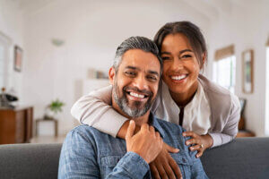 Why Personal Relationships Matter in Financial Planning- Smiling couple sitting together in a cozy home setting. The man, with salt-and-pepper hair and a beard, is seated in front, while the woman stands behind him with her arms affectionately wrapped around his shoulders. Both are dressed casually and radiate warmth and happiness, creating a relaxed and intimate atmosphere.